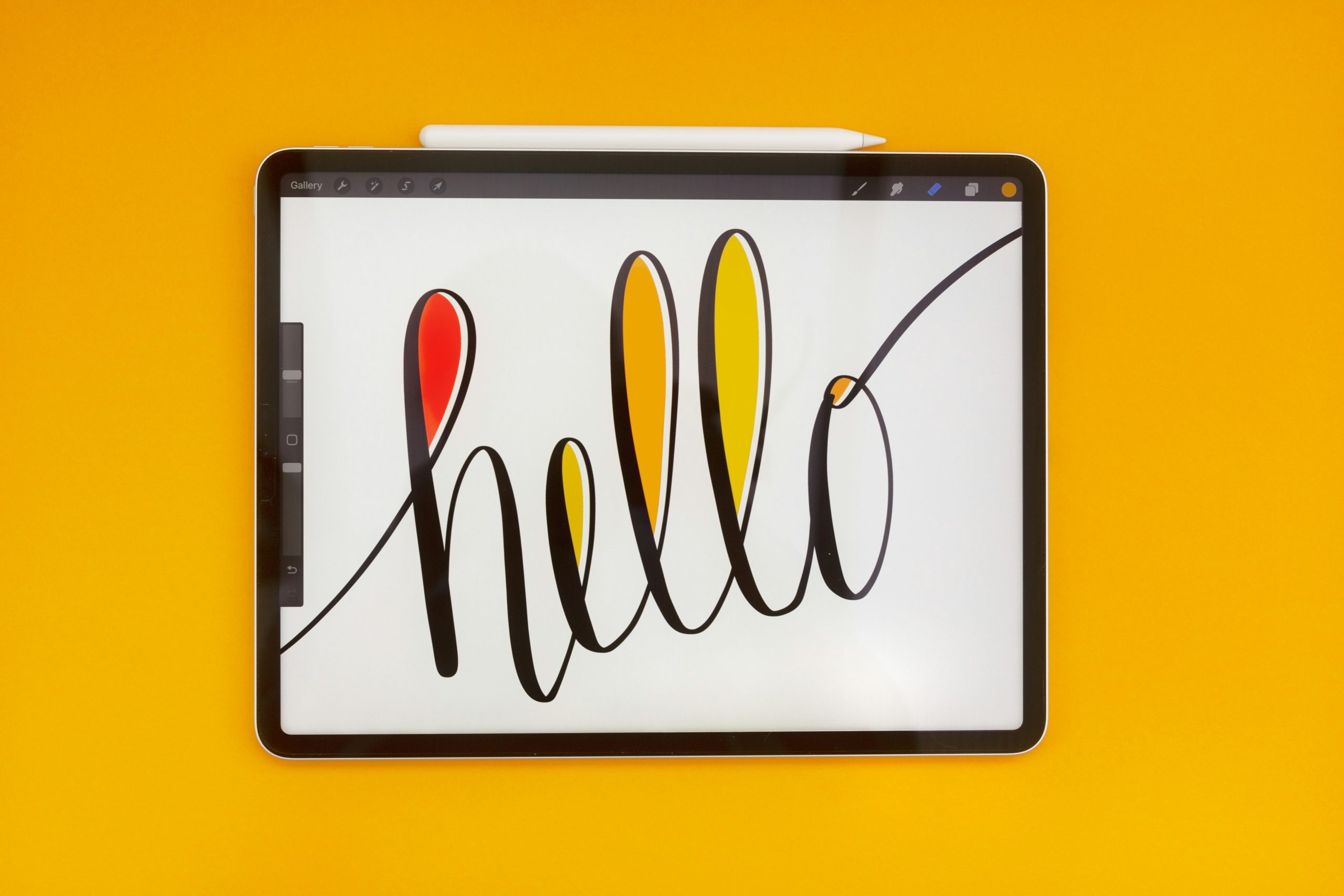 An iPad Pro with the word 'hello' hand lettered on screen. The iPad is set against a bright orange background. Attached to the top of the iPad is an Apple pencil