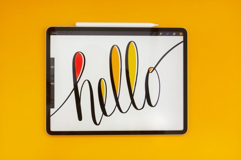 An iPad Pro with the word 'hello' hand lettered on screen. The iPad is set against a bright orange background. Attached to the top of the iPad is an Apple pencil